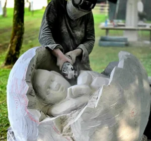 Raphaëlle Duval en train de sculpter une œuvre représentant un fœtus lové au cœur d’une Terre Mère qui éclate, vue de près et de haut, réalisée lors du symposium de sculpture en Italie en 2025, dimensions 100 × 70 × 70 cm.